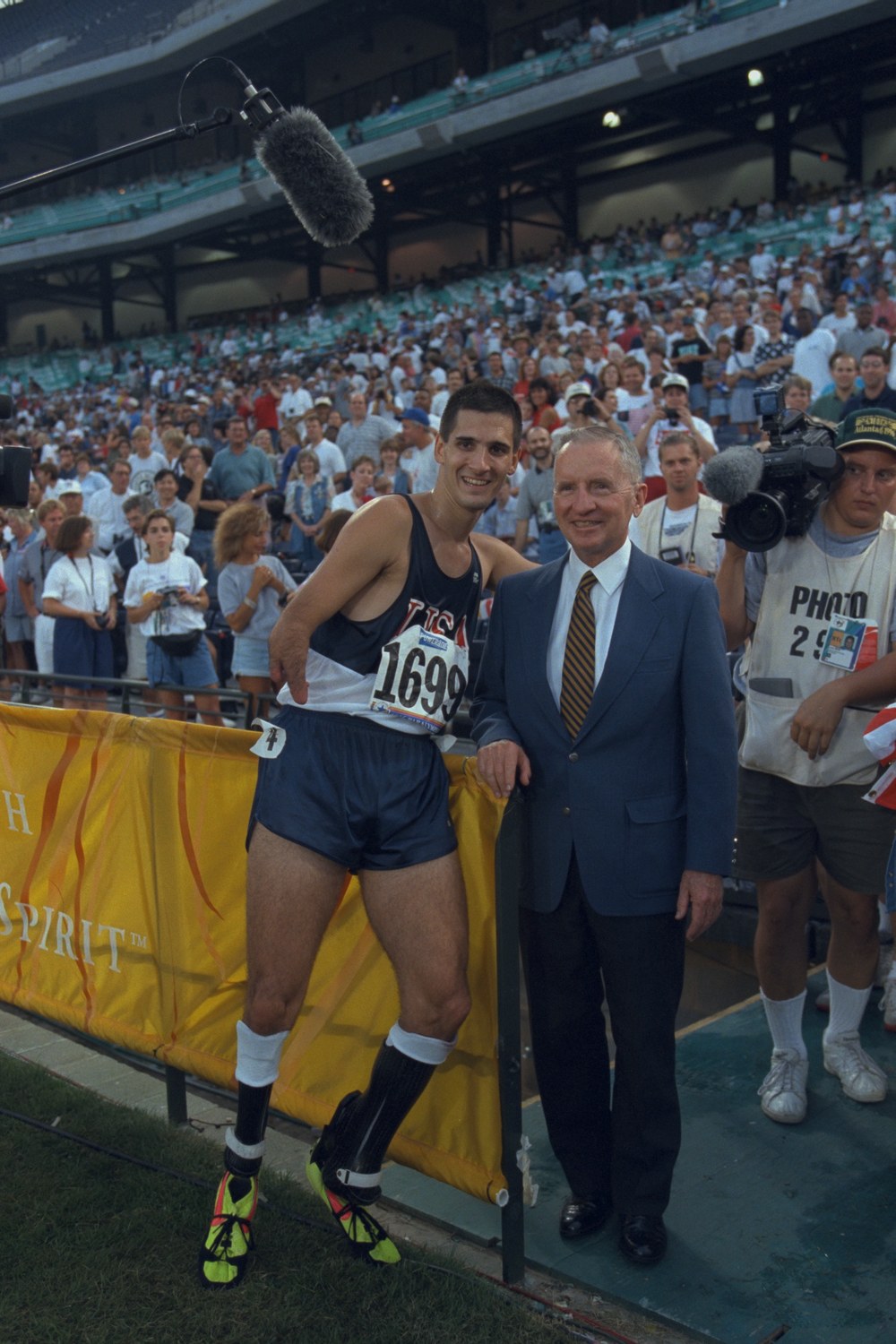 Tony Volpentest in USA competition kit with bib 1699, running blades visible, standing at a barrier beside Ross Perot in a suit; camera crew and a full crowd behind them
