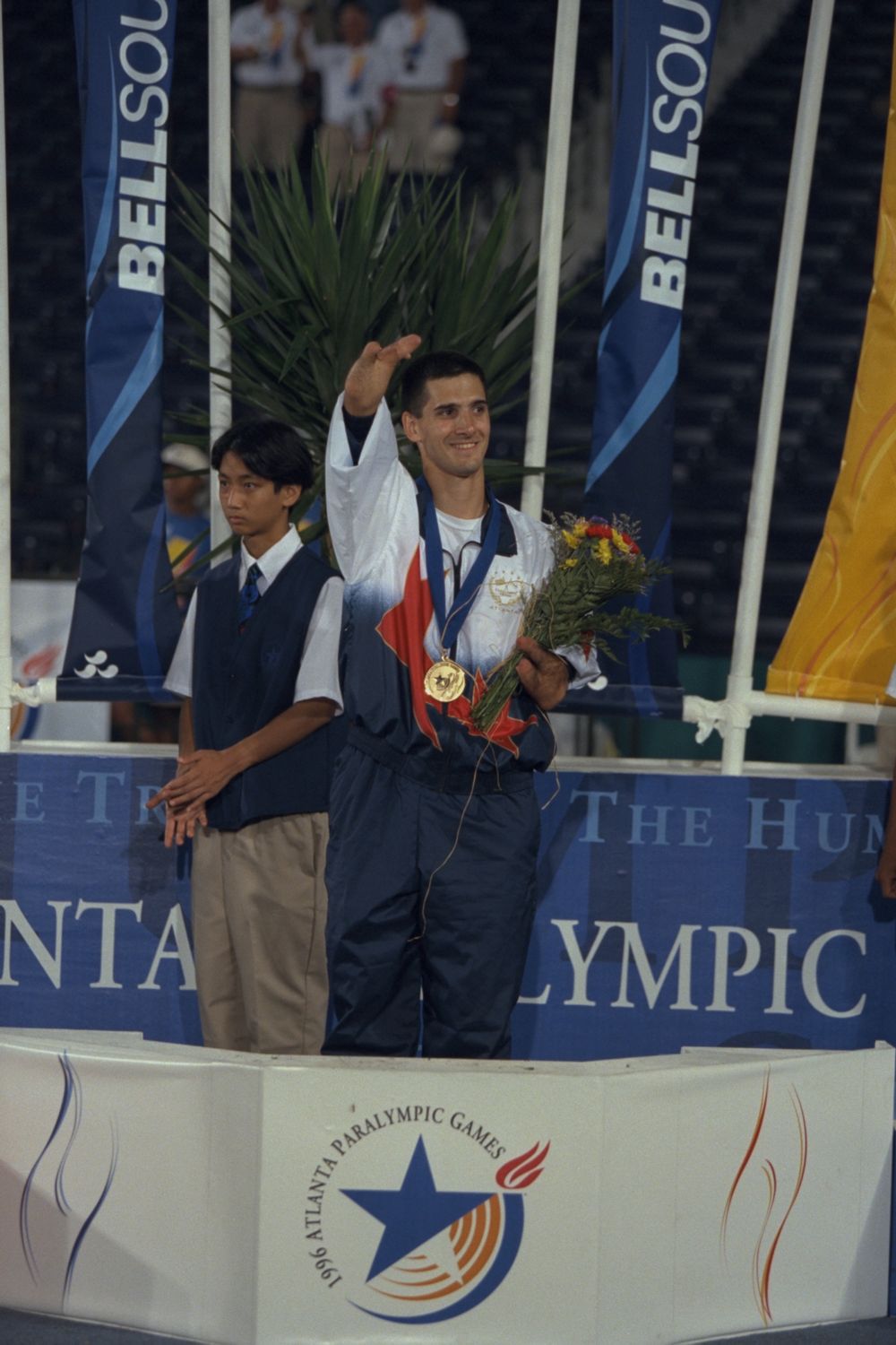 Tony Volpentest waving from the podium, gold medal and bouquet, a uniformed attendant standing behind