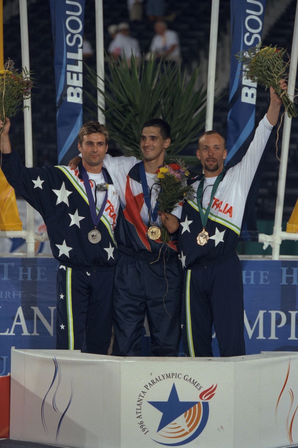 Three medalists on the podium, Tony Volpentest in the center, one arm around the silver medalist and a bouquet in the other