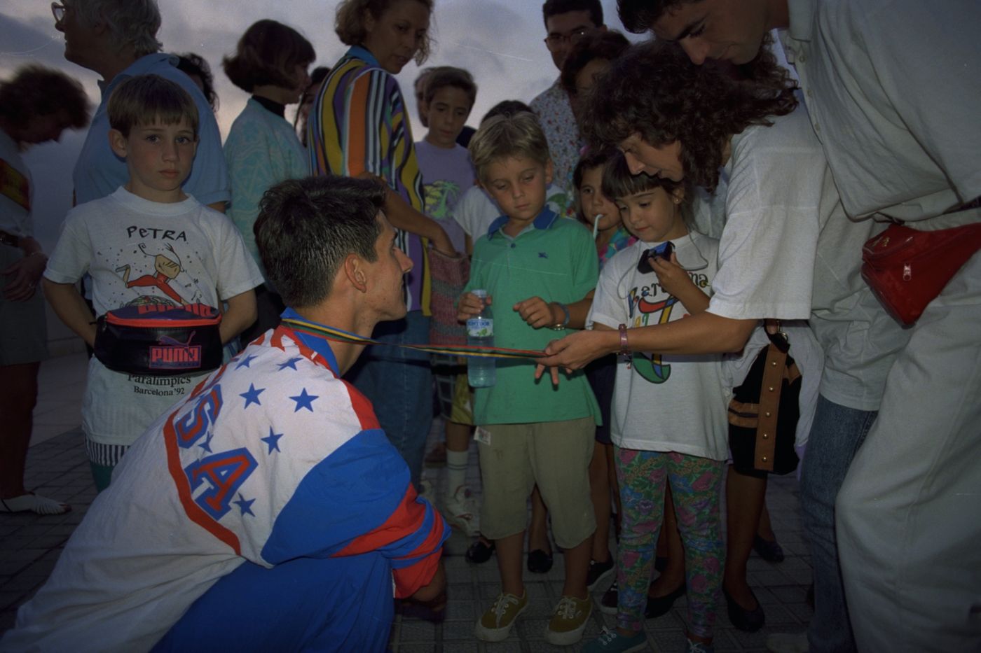 Tony Volpentest kneeling in a ring of children, holding out his medal on its ribbon, Barcelona 1992