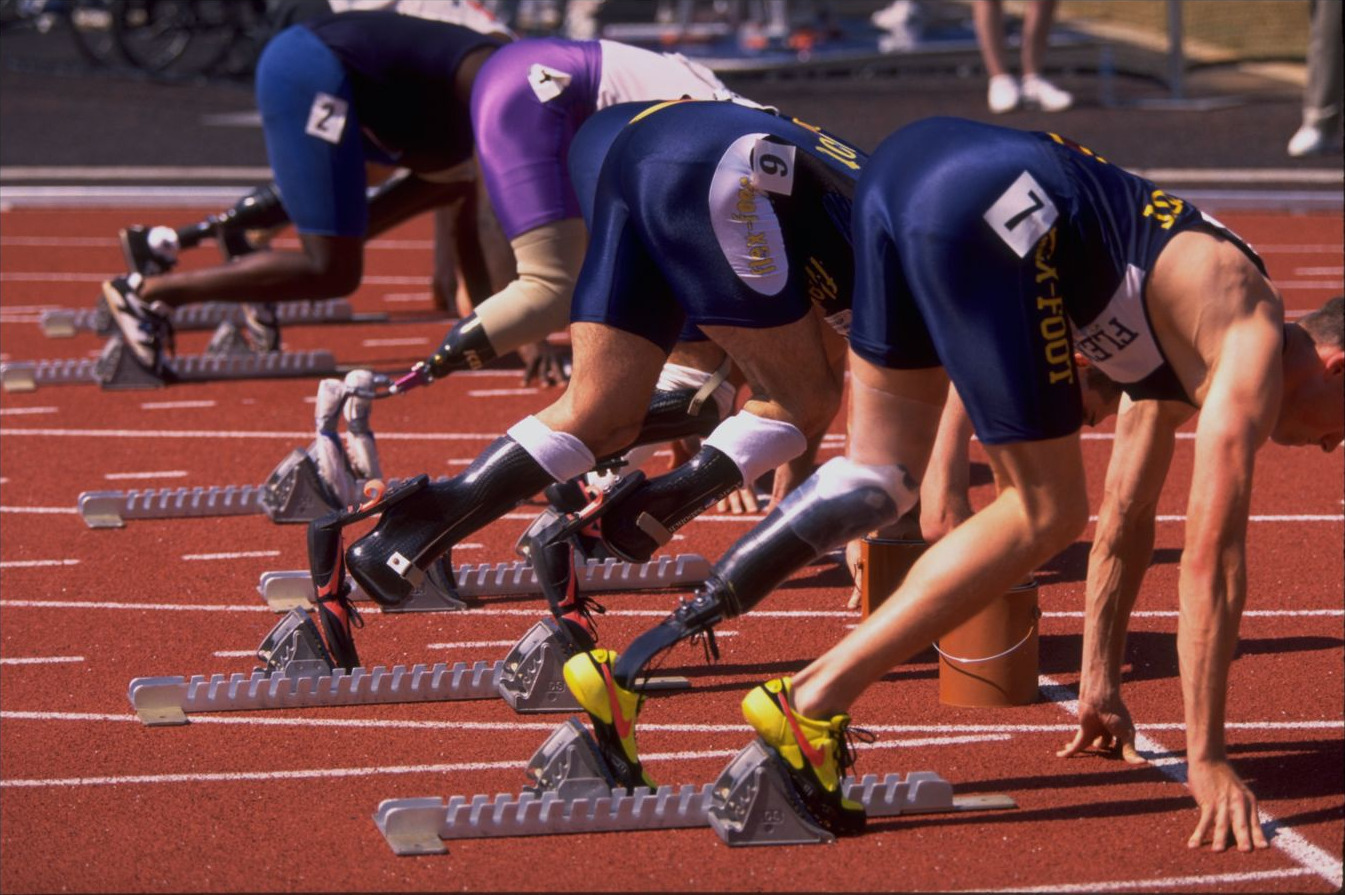 A row of sprinters down in the blocks seen from the side, carbon-fiber blades braced against the pedals